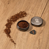 Aluminum Storage Puck alongside a guitar pick, featuring a metal container with a pile of brown shavings, illustrating its compact and discreet design.