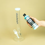 Hand holding a 12oz Black Label Bong Cleaner bottle next to a glass bong, highlighting its use for effective cleaning of smoking accessories.